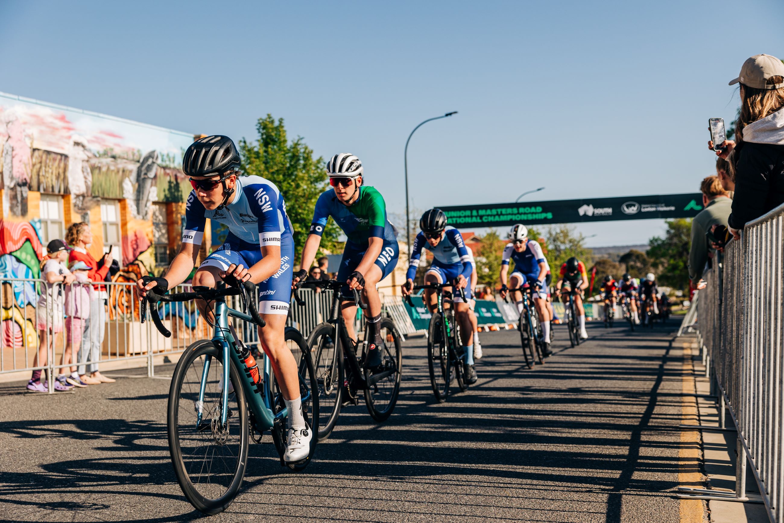 A New South Wales NSW rider leads the peloton under the finish arch at the 2024 AusCycling Masters and Junior Road National Championships criterium in Loxton