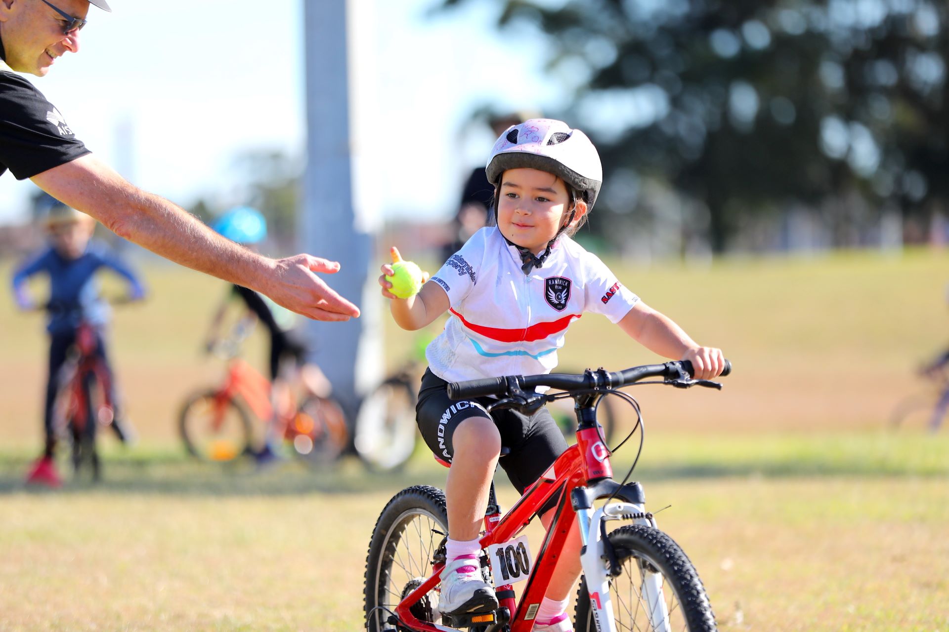 A child riding a bike holding a tennis ball during a training exercise in an AusBike coaching session