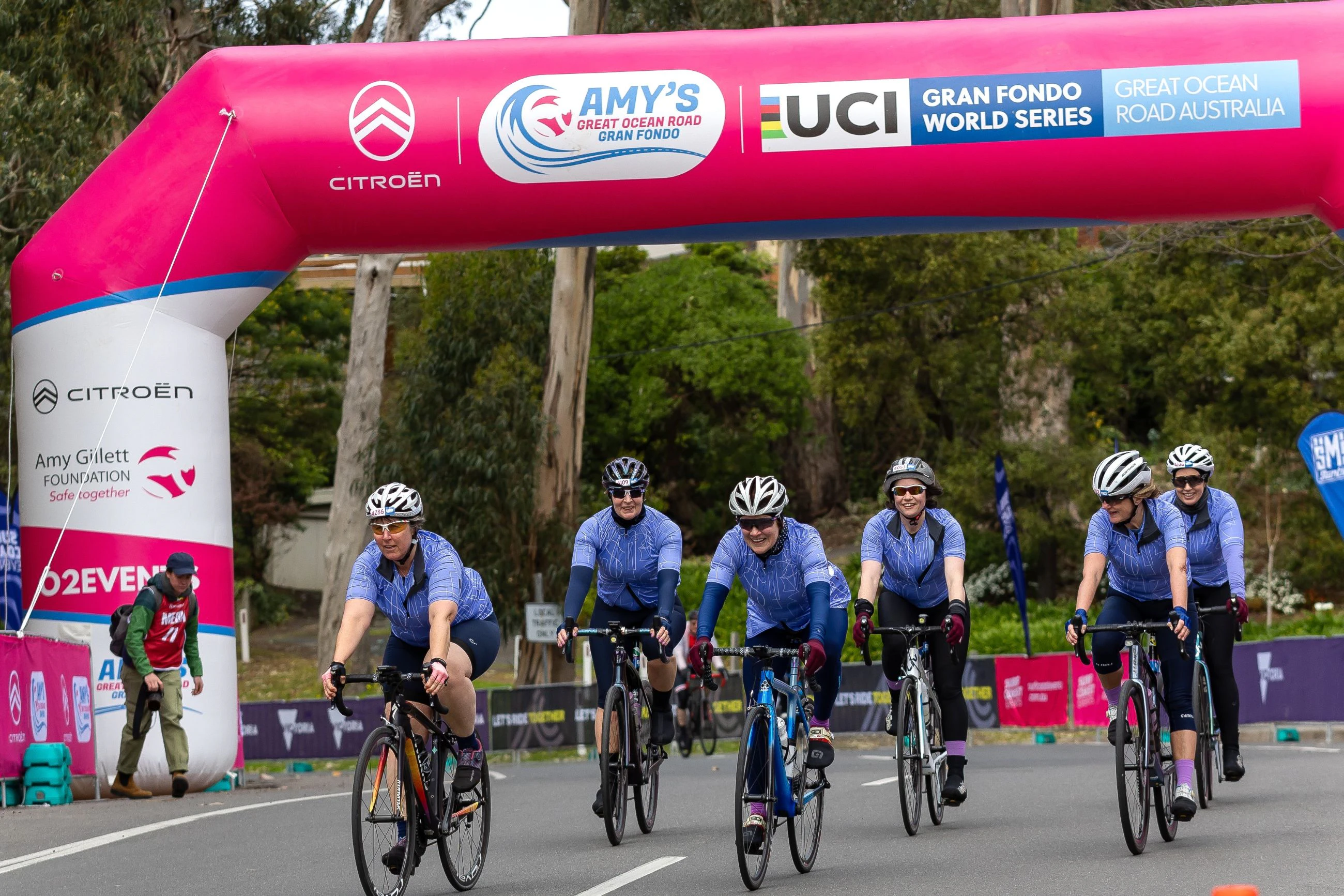 A group of female cyclists finish the Amy's Gran Fondo, crossing under a large pink inflatable arch with smiles on their faces. Photo by Con Chronis.