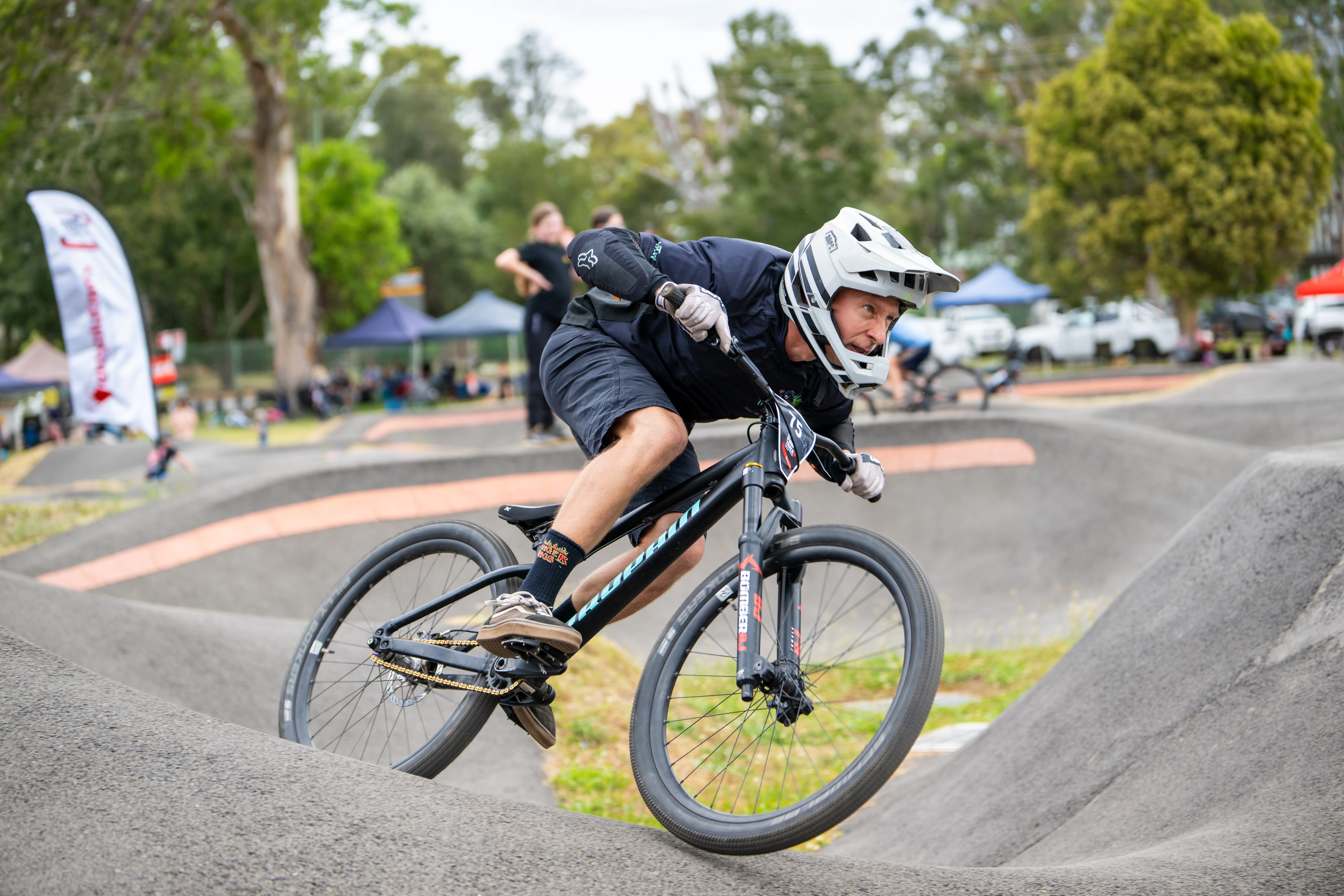 Cyclist wearing a white full-face helmet leans into a banked turn on an asphalt pump track, with spectators and event tents in the background.