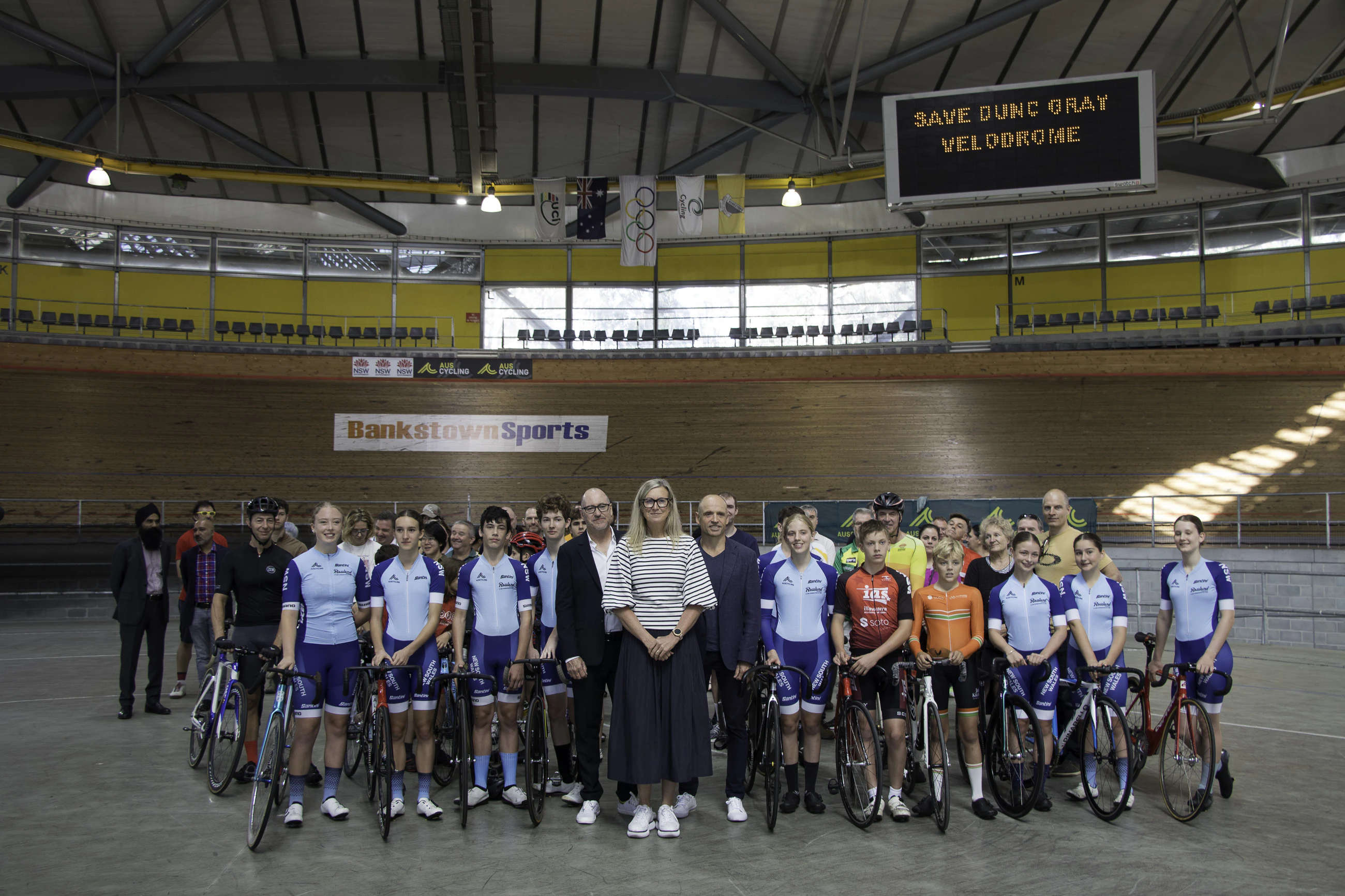 Marne Fechner, CEO of AusCycling; Mark Arbib, CEO of the Australian Olympic Committee (AOC); and Olympic games track cycling gold medallist Scott McGrory pose with members of the Sydney and New South Wales cycling community and Canterbury-Bankstown local sporting community in Dunc Gray Velodrome with the message "Save Dunc Gray Velodrome" on the big screen behind them.