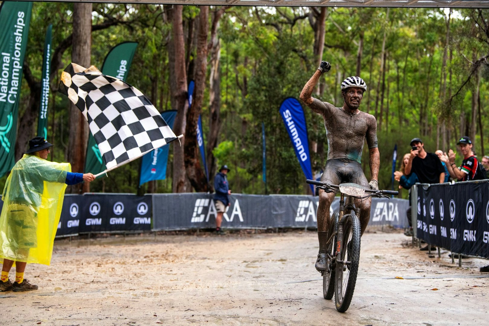 Covered in mud so as to be almost indistinguishable, mountain bike rider Cameron Ivory crosses the finish line with a fist pump at the 2024 XCO National Championships