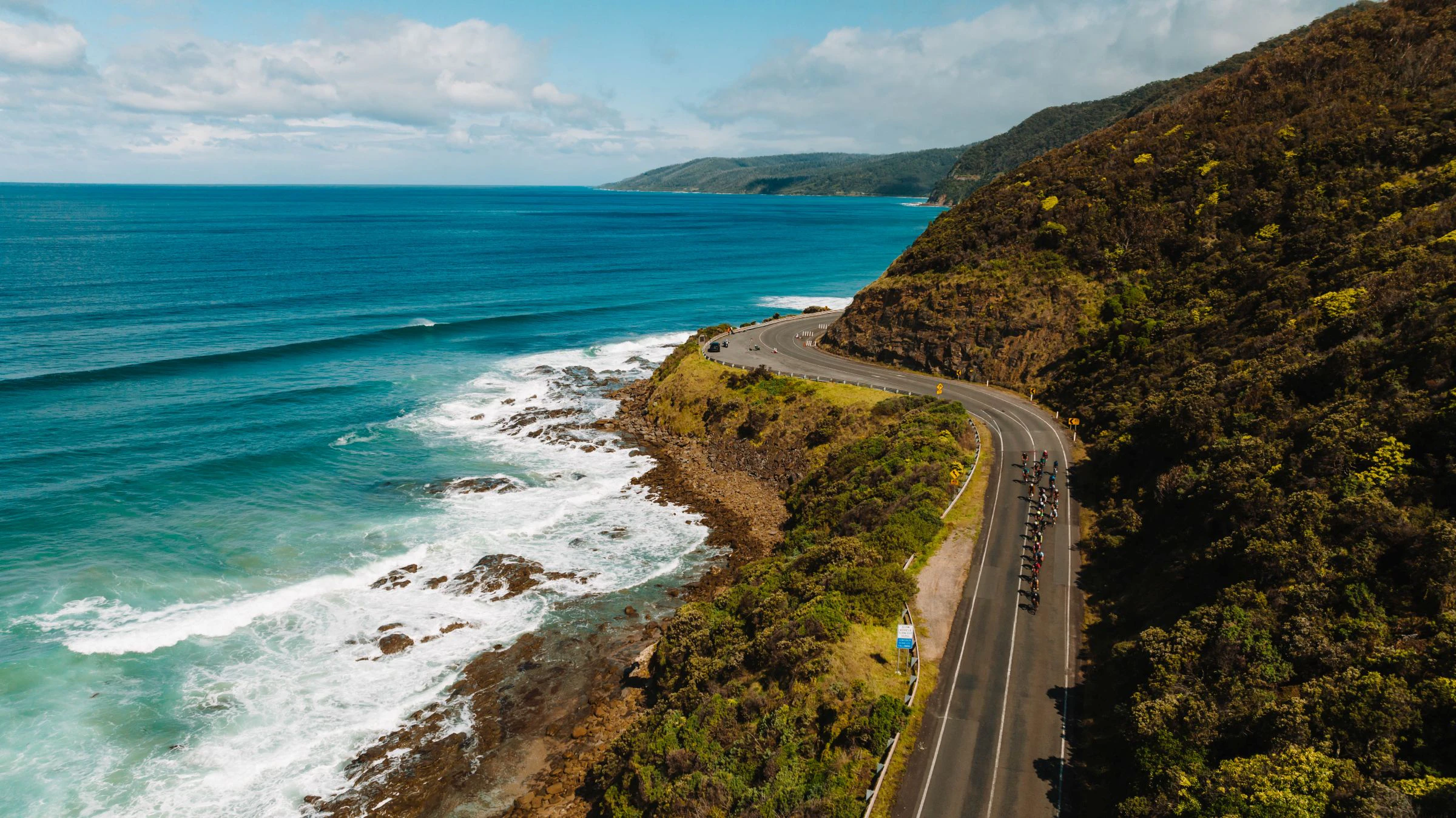 Aerial view of cyclists following a coastal bend on the Great Ocean Road during Amy’s Gran Fondo, with ocean waves below and wooded hills beside the road.