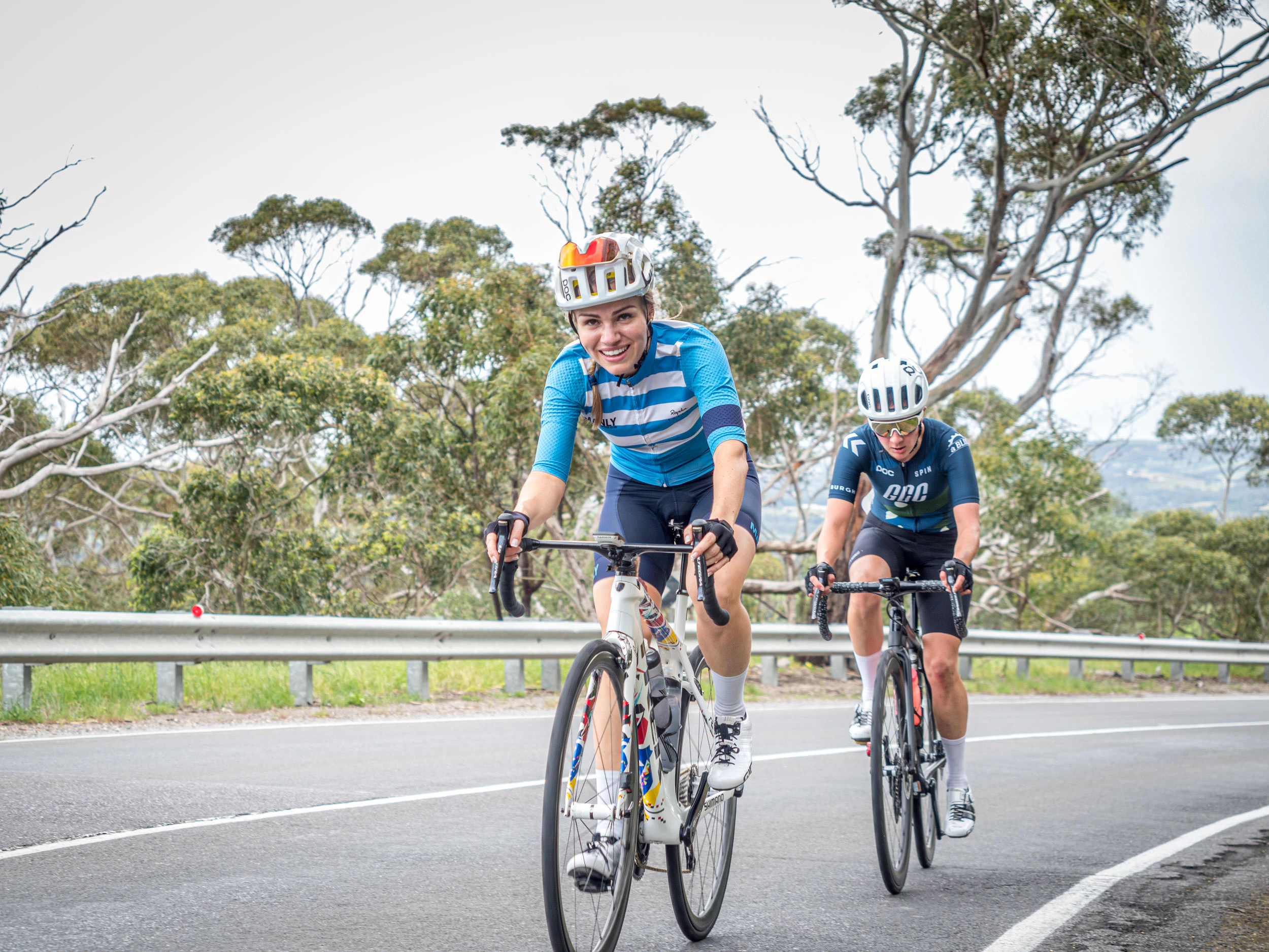 Two cyclists riding road bikes on a tree-lined road, with the rider in front smiling towards the camera.