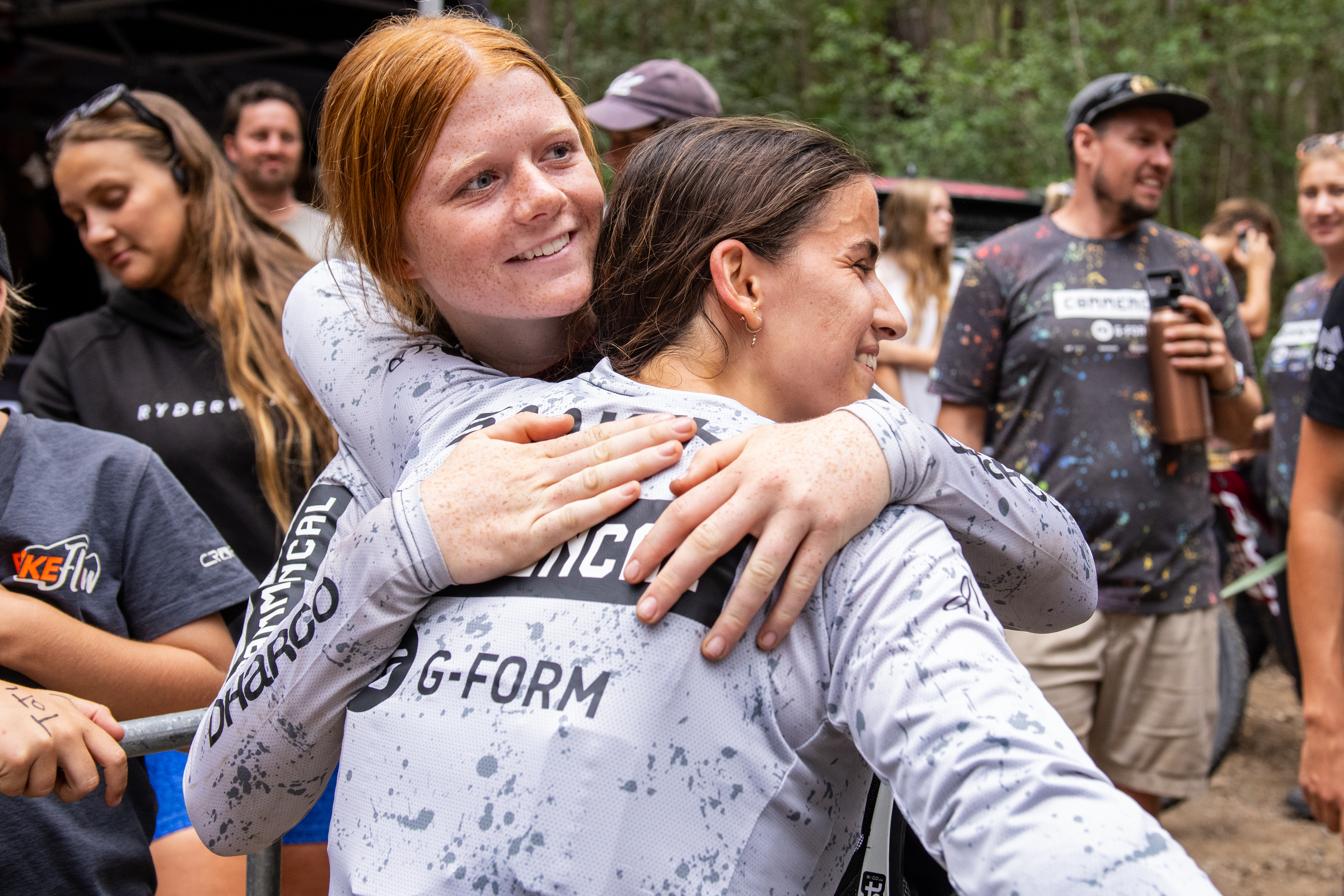 Two smiling mountain bike riders in matching grey jerseys hugging, with other riders and spectators gathered behind them.
