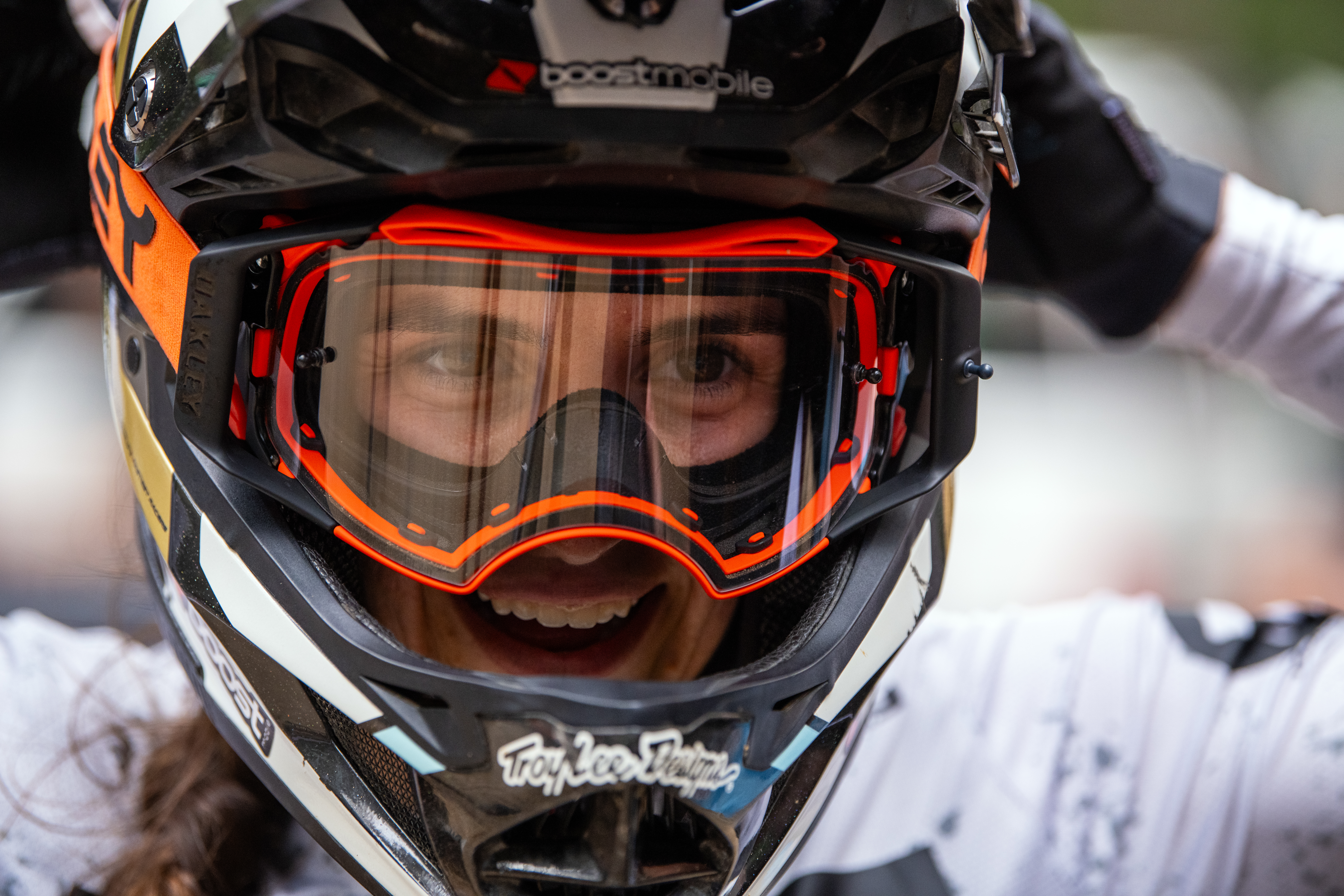 Close-up of a smiling mountain bike rider wearing orange goggles and a full-face helmet, with gloved hands raised to the helmet.