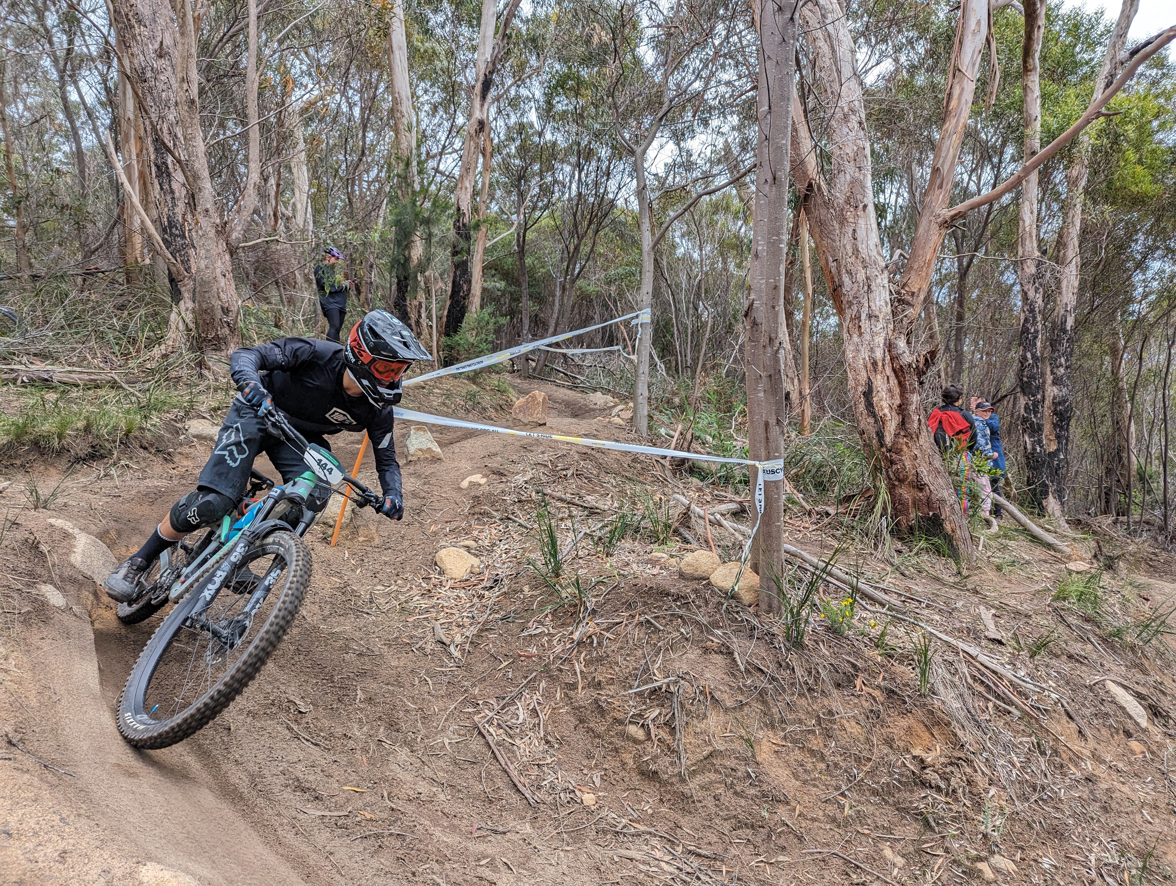 Enduro mountain bike rider in black kit leaning into a rocky corner on a taped course through eucalyptus forest.