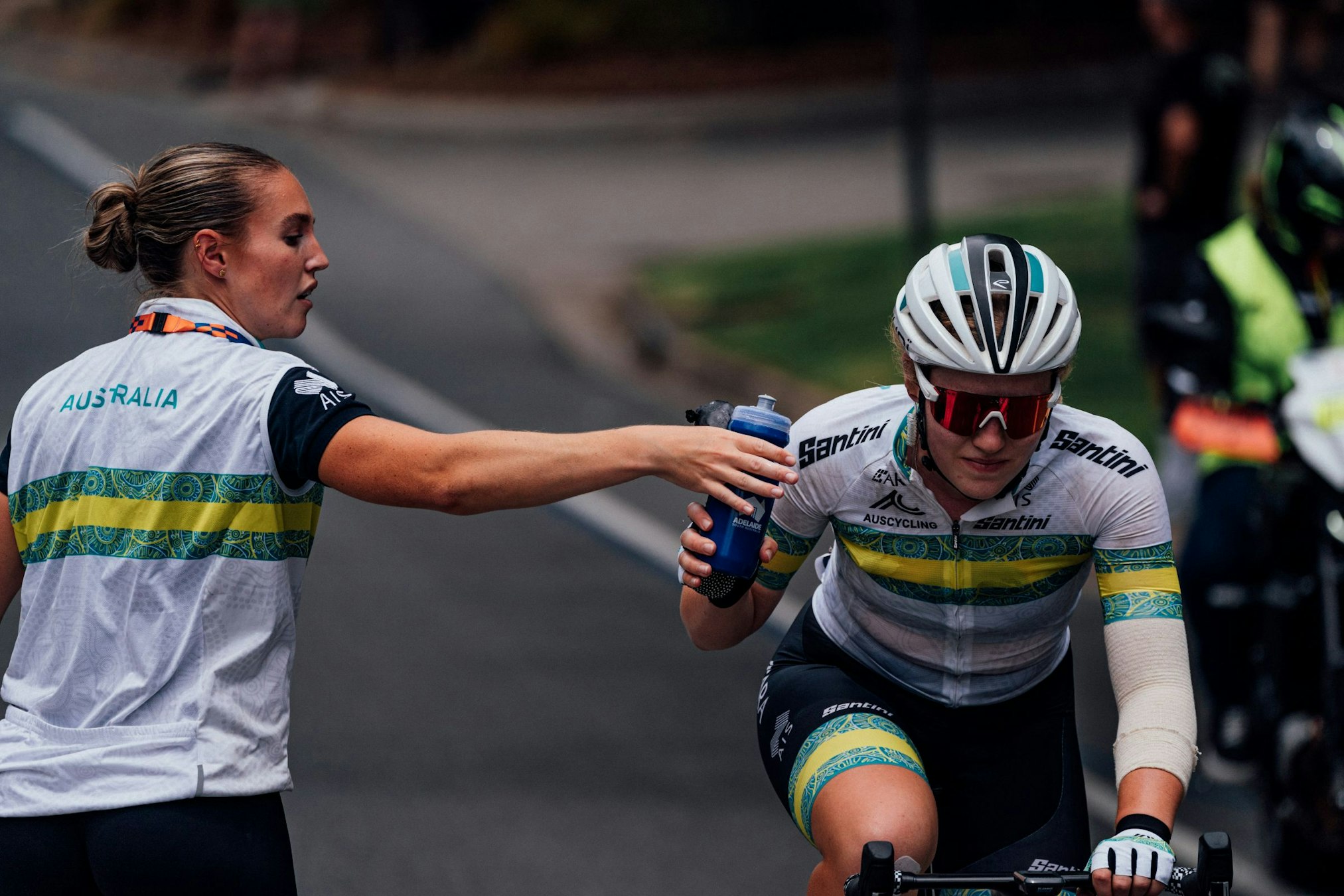 ARA Australian Cycling Team rider Lauren Bates takes a bottle from the feed zone soigneur during the 2025 Santos Tour Down Under women's race