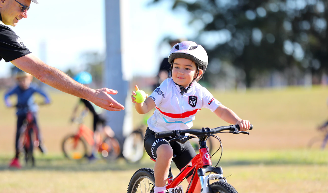 AusBike student wearing white jersey and helmet passing tennis ball while riding