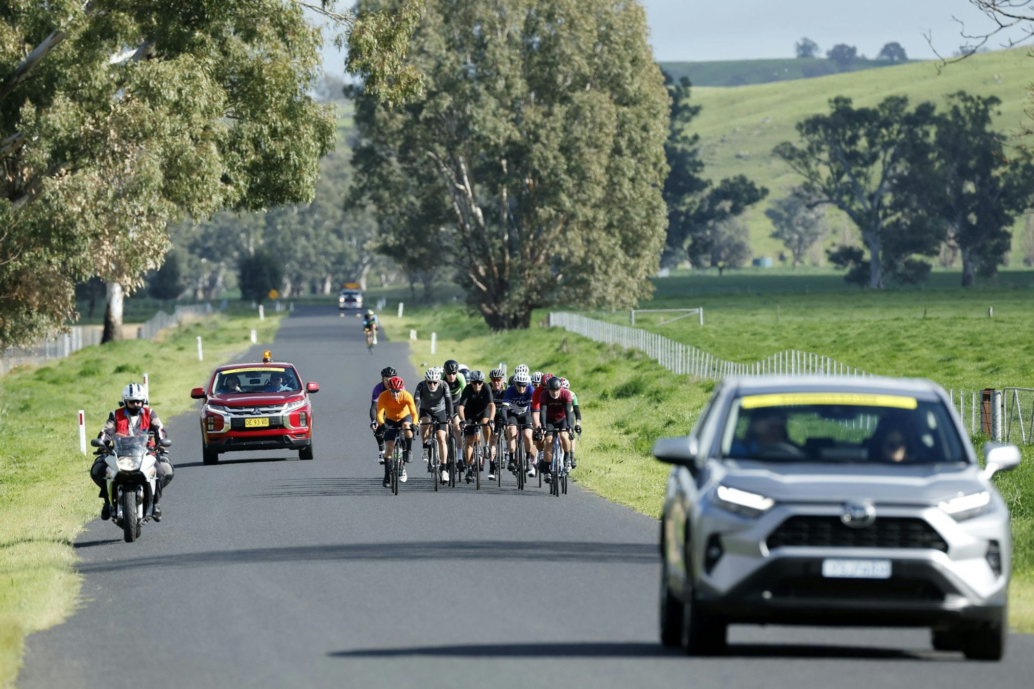 A bunch of road racing cyclists shepherded by a lead vehicle, follow car and motorcycle on a closed road during the 2025 AusCycling Masters Road National Championships in Wagga Wagga. Con Chronis