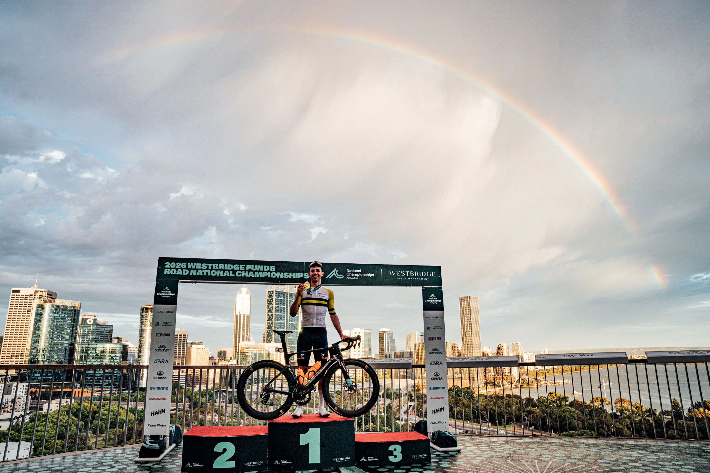 Patrick Eddy of team Brennan, Australian elite men road race champion, poses with bicycle on the podium of the Westbridge Funds Road National Championships in Perth with a rainbow arching above him and the Perth skyline behind.