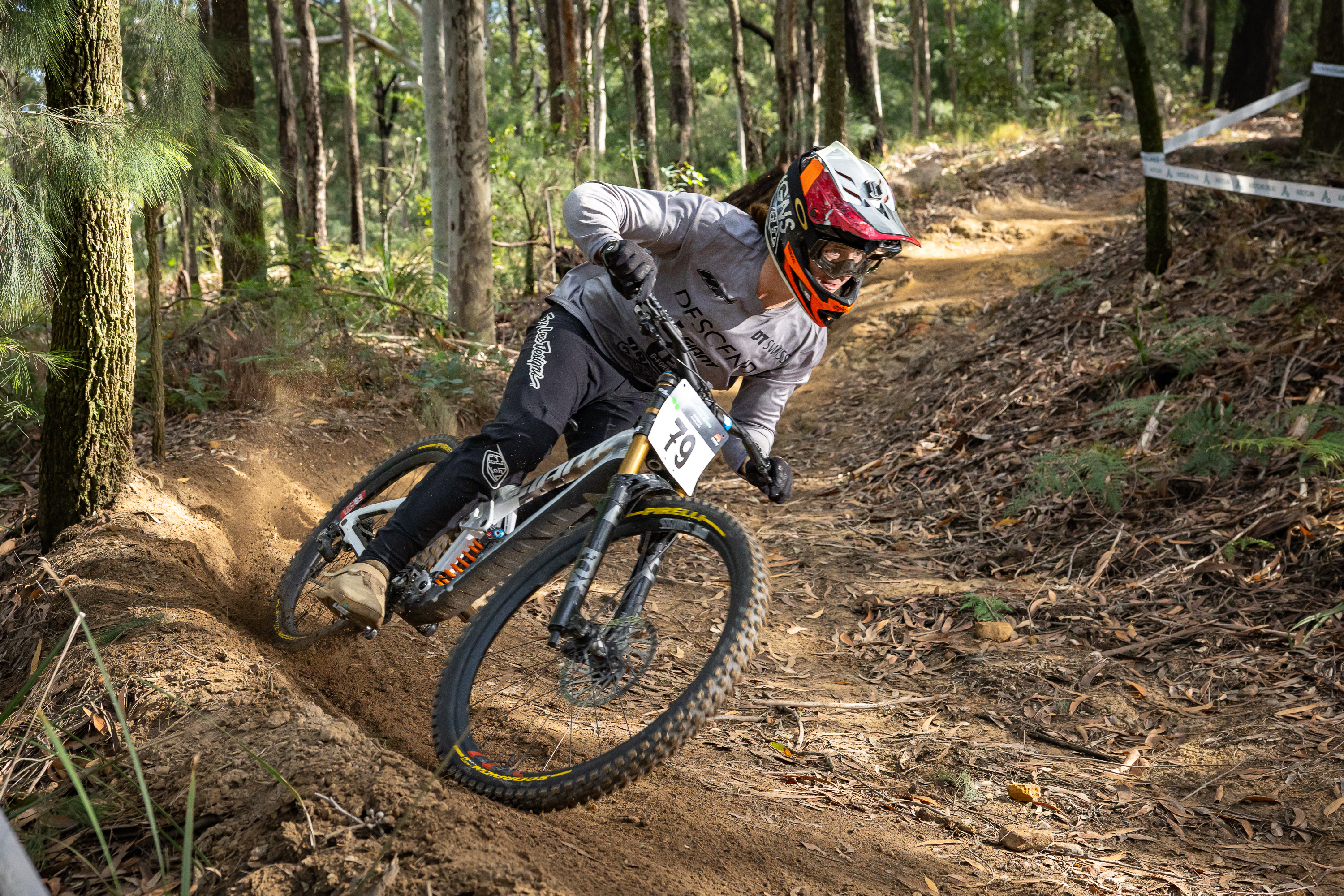 Mountain bike rider in a grey jersey and full-face helmet leaning into a dirt corner, throwing up dust on a forest race course.